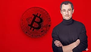 Eine Bitcoin-Münze vor einem roten Hintergrund. Rechts am Bild steht ein Mann mit schwarzem Rollkragenpullover.