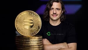 Ein Mann mit schulterlangen dunklen Haaren und Schnurrbart steht mit verschränkten Armen neben einem Stapel goldener Solana-Coins. Er trägt ein schwarzes T-Shirt mit grünem „Greenfield“-Logo. Im Vordergrund steht eine große Solana-Münze auf gestapelten Coins, während der Hintergrund dunkel mit violett-blauen Lichteffekten ist.