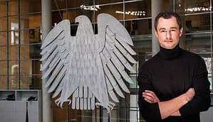 Der Deutsche Bundesadler hängt im Bundestag und rechts sieht man einen Mann mit schwarzem Rollkragenpullover