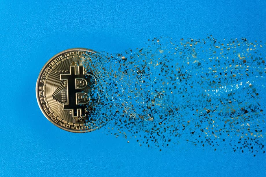 Vor einem blauen Hintergrund sieht man eine Bitcoin-Münze, die sich in viele kleine Einzelteile auflöst