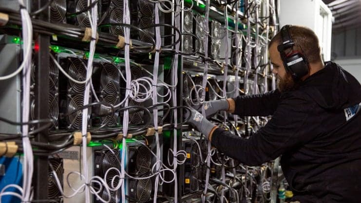 Ein Mann mit Kopfhörern und Handschuhen arbeitet an einer großen Reihe von Bitcoin-Mining-Maschinen mit zahlreichen Kabeln in einer industriellen Umgebung.