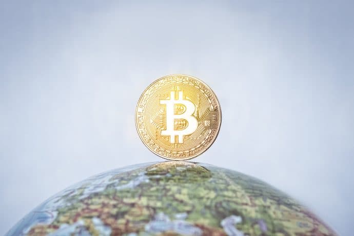 Man sieht einen Globus auf dem eine Bitcoin-Münze steht vor blauem Hintergrund.