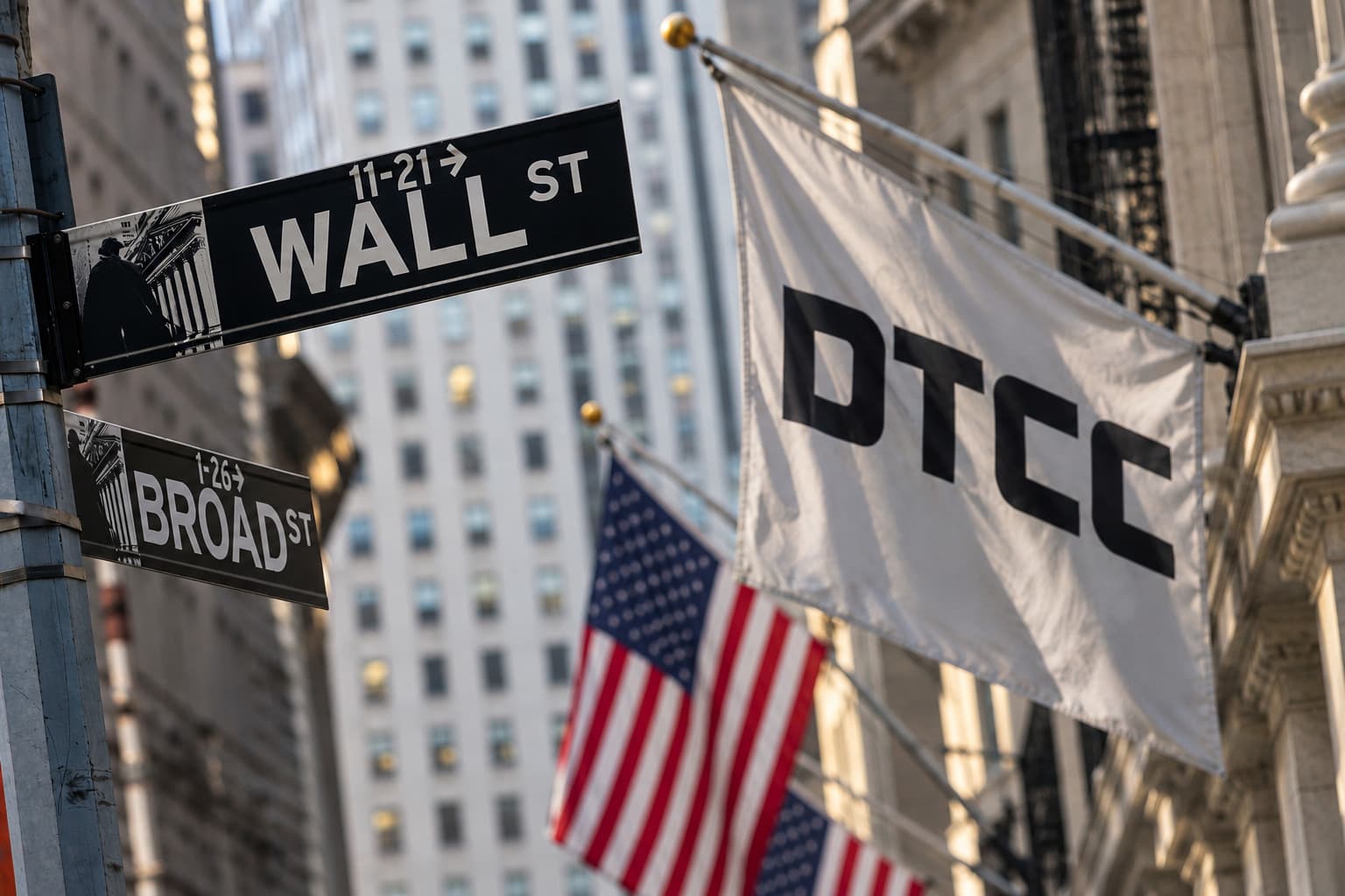 Wall-Street-Straßenschild in New York neben einer weißen Flagge mit DTCC-Logo und US-amerikanischen Flaggen im Hintergrund.
