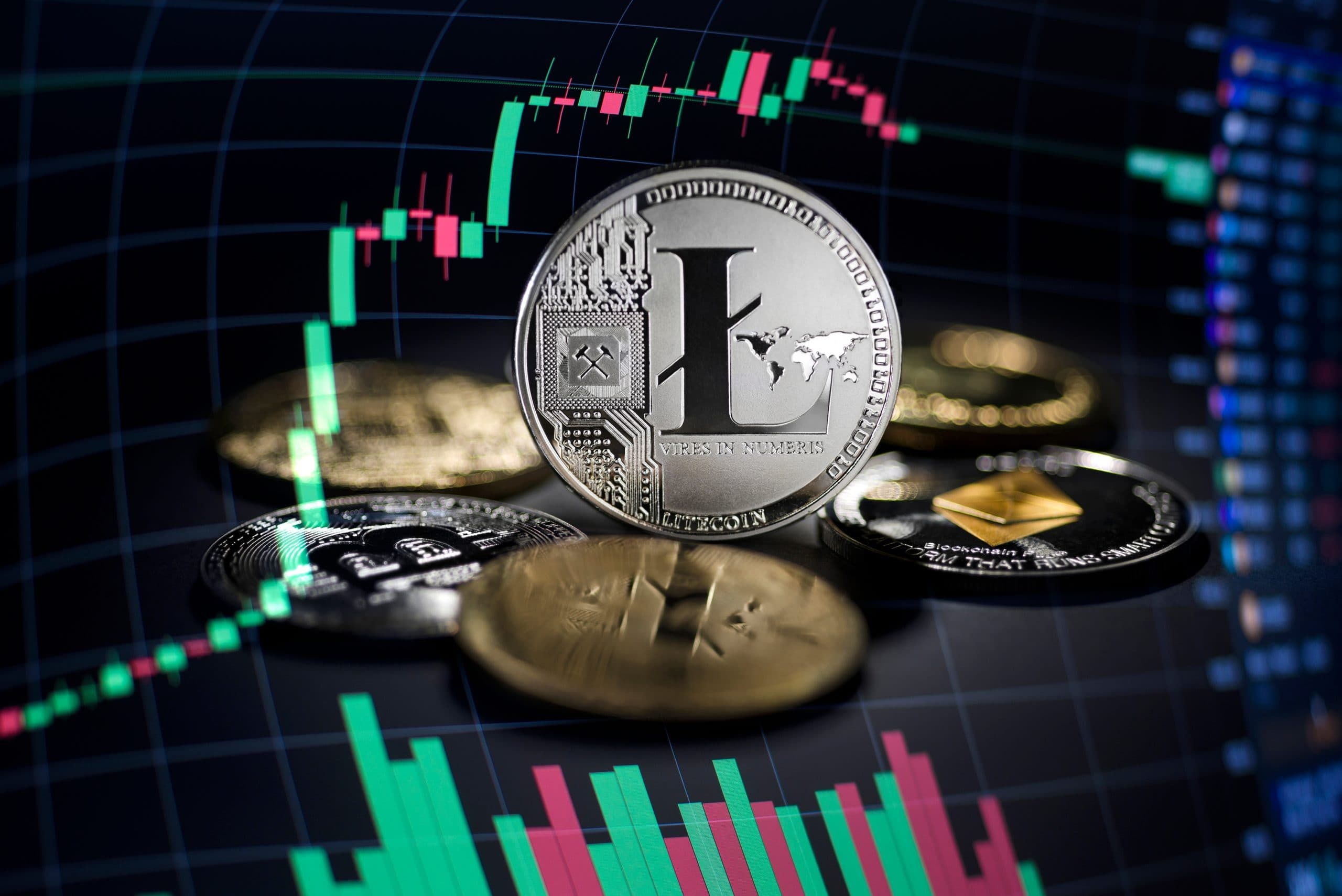 Ein Stapel verschiedener Kryptowährungsmünzen, darunter auch Litecoin, liegt vor einem digitalen Handelschart mit Kerzenständern und Linien im Hintergrund.