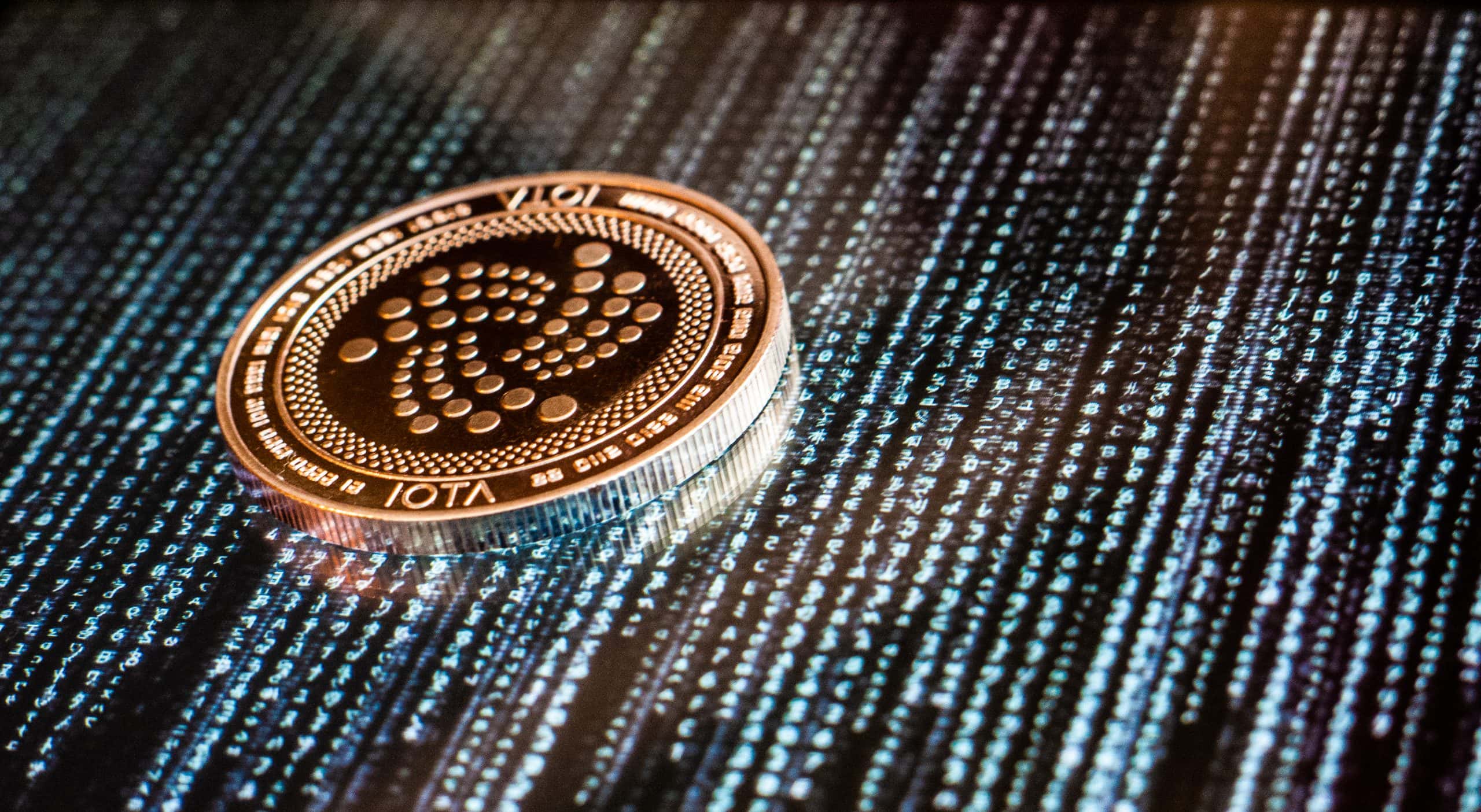 Eine physische IOTA-Münze ruht auf einer Oberfläche, die einen digitalen Binärcode und alphanumerische Zeichen anzeigt.