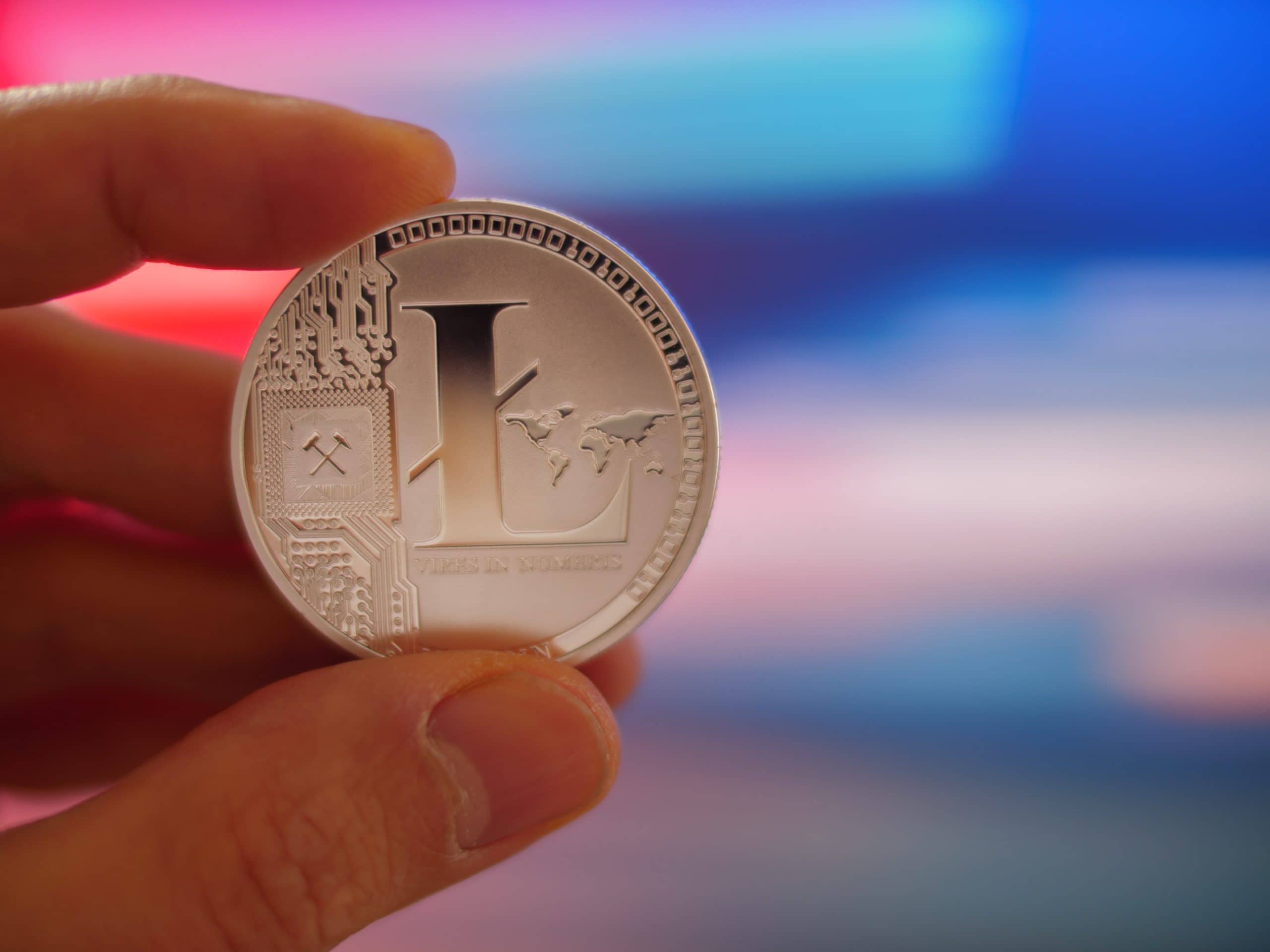 Eine Hand hält eine silberne Litecoin-Münze mit dem charakteristischen digitalen Schaltkreisdesign und der Kartengravur vor einem unscharfen bunten Hintergrund.