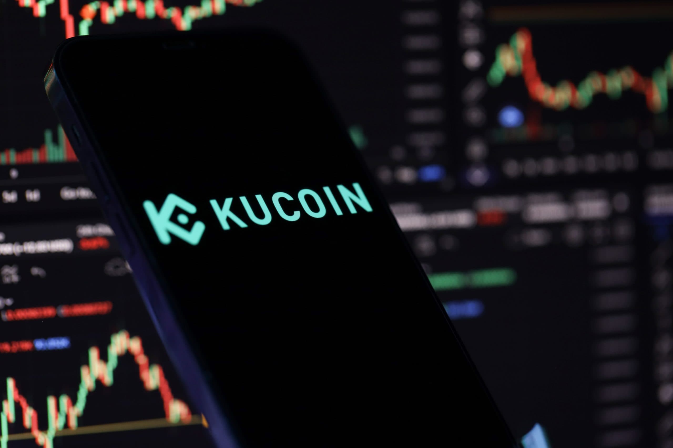 Smartphone mit Kucoin Emblem vor dunklem Hintergrund mit Graphen