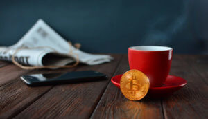 bitcoin-münze-vor-einer-kaffeetasse-auf-einem-holztisch-mit-zeitung-im-hintergrund