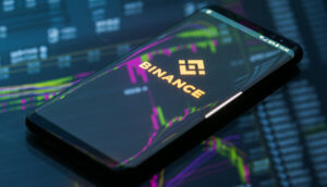 Smartphone, das das Logo von Binance zeigt, liegt auf einem gläsernen Grund, auf dem sich eine Chart spiegelt.