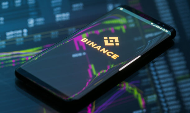 Everipedia: Wikipedia-Alternative nun auf Binance verfügbar Smartphone, das das Logo von Binance zeigt, liegt auf einem gläsernen Grund, auf dem sich eine Chart spiegelt.