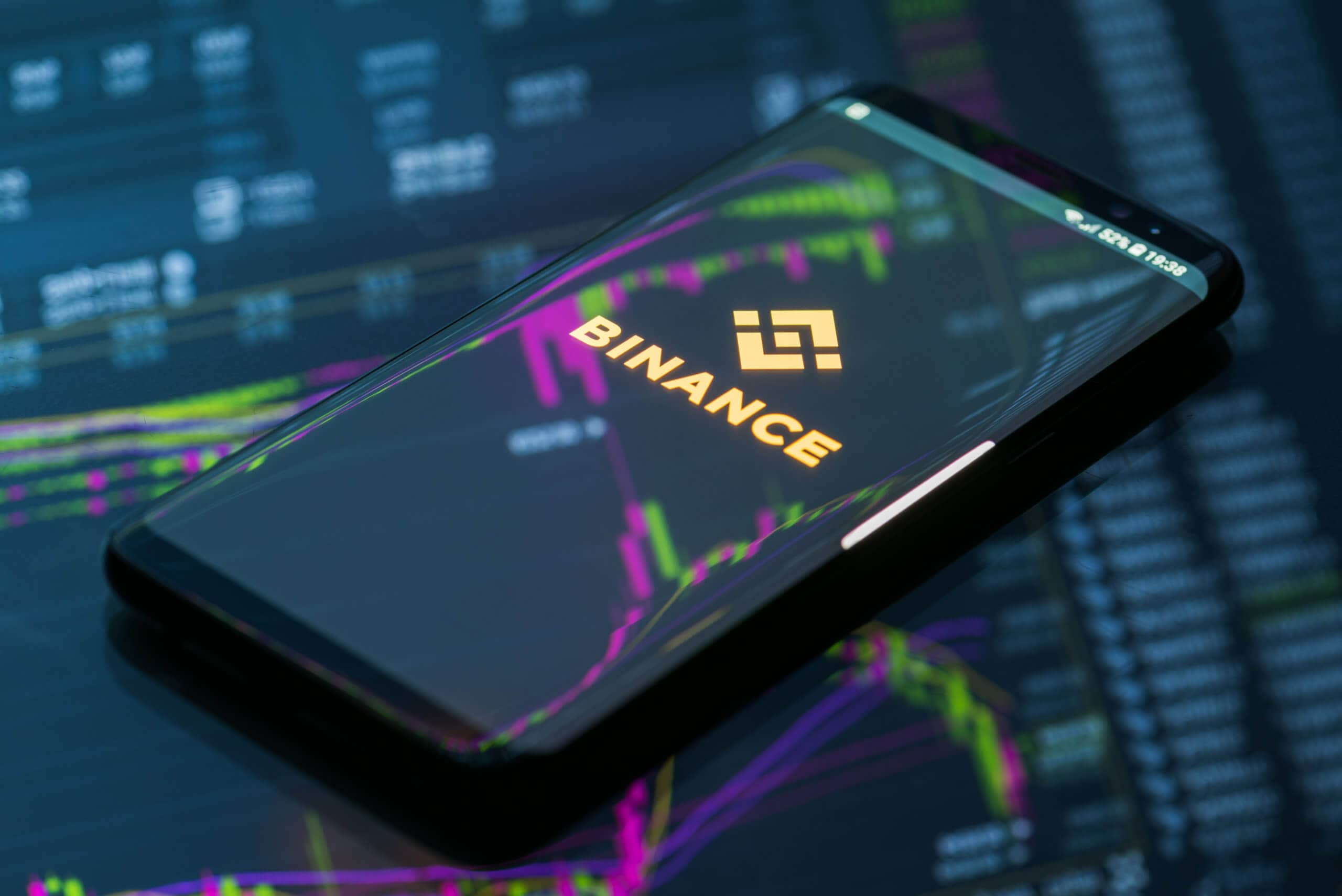 Smartphone, das das Logo von Binance zeigt, liegt auf einem gläsernen Grund, auf dem sich eine Chart spiegelt.