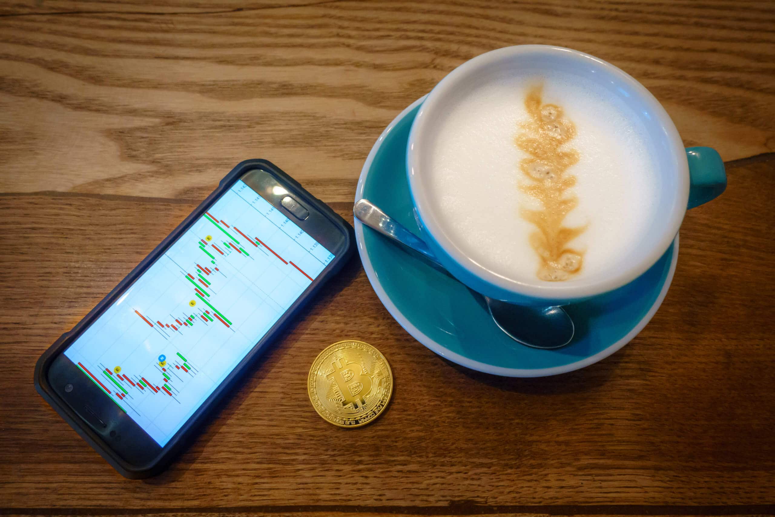 Smartphone das Charts zeigt, neben einer goldenen Bitcoin-Münze und einer Kaffeetasse mit Latteart auf einem braunen Holztisch