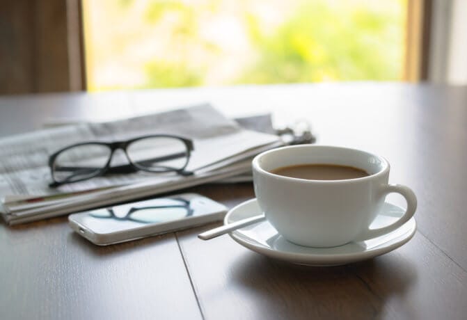 Tasse Kaffe auf dem Tisch mit einem Fenster im Hintergrund. Neben der Tase ein Smartphone und eine Zeitung, auf der eine zugeklappte Brille liegt