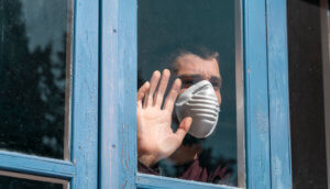 Sehnsuchtsvoll aus dem Fenster blickender Mann mit Mundschutz