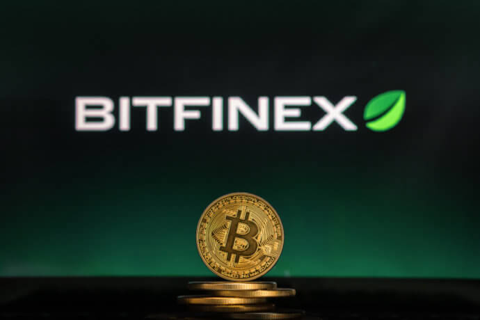 In der Mitte steht eine goldene Bitcoin Münze und im Hintergrund wird die Bitfinex Börse angezeigt