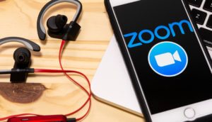 smartphone, auf dem das zoom-logo erkennbar ist, liegt neben kopfhörern auf einem holztisch