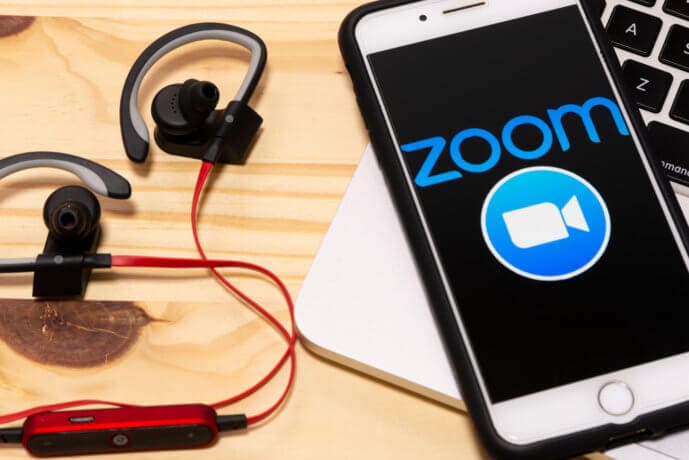 smartphone, auf dem das zoom-logo erkennbar ist, liegt neben kopfhörern auf einem holztisch