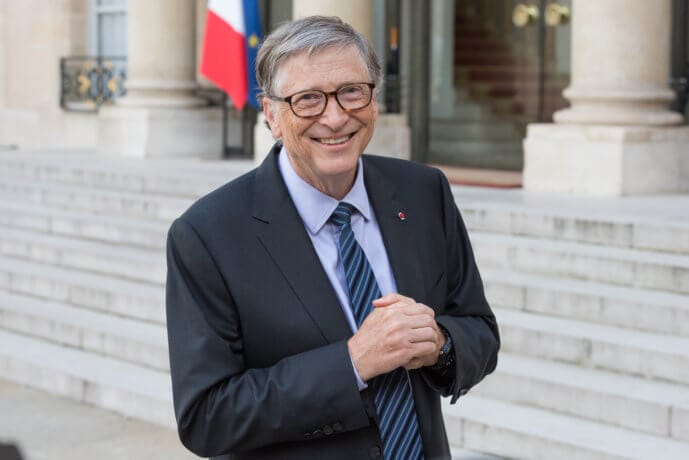 bill gates steht im anzug mit blau schwarzer krawatte und grauem haar vor einem gebäude, von dem man die treppenstufen sieht und lächelt