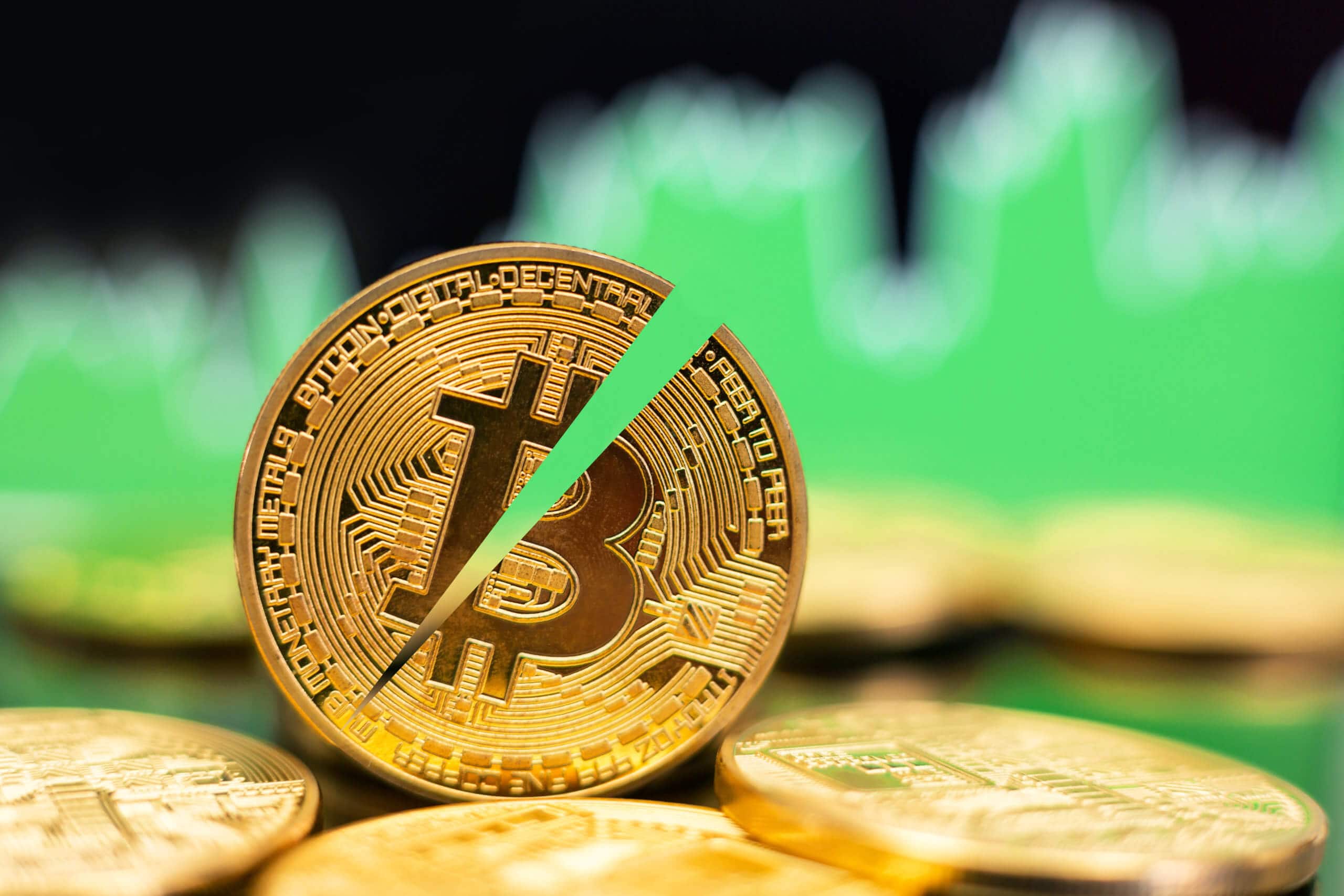 halbierte bitcoin münze die zwischen liegenden bitcoin münzen steht, im Hintergrund grüne Charts als Symbol für einen Bitcoin-Kurs-Anstieg nach dem Halving