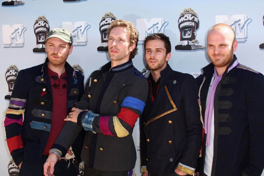 Zumo: Coldplay Bassist unterstützt Bitcoin App Die Bandmitglieder von Coldplay stehen vor einer Plakatwand für Fotoshooting.