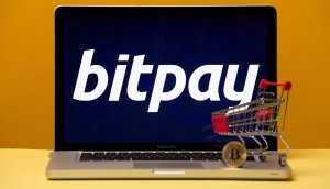 Laptop mit Bitpay-Logo auf dem Bildschirm und Miniatur-Einkaufswagen samt Bitcoin-Münue