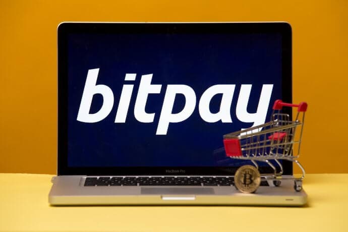 Laptop mit Bitpay-Logo auf dem Bildschirm und Miniatur-Einkaufswagen samt Bitcoin-Münue