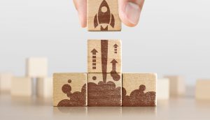 Unternehmensgründung, Start, neues Projekt oder neues Ideenkonzept. Hölzerne Blöcke mit Raketengrafik in Pyramidenform angeordnet und ein Mann hält den oberen.