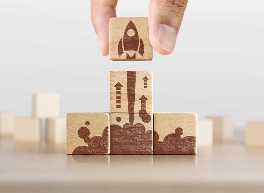Blockrockets wählt AZHOS zum Blockchain-Start-up des Monats Unternehmensgründung, Start, neues Projekt oder neues Ideenkonzept. Hölzerne Blöcke mit Raketengrafik in Pyramidenform angeordnet und ein Mann hält den oberen.