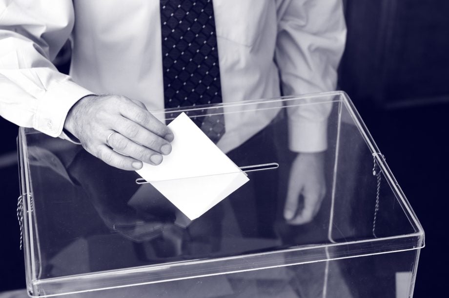 Aave: DeFi-Plattform mit neuer Governance auf Ethereum-Mainnet Ein Mann in Hemd und Krawatte steckt einen Zettel in eine transparente Wahlurne.