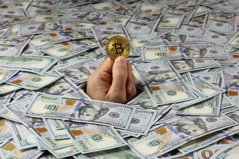MicroStrategy könnte mit Bitcoin Dump 3-fachen Jahresprofit einfahren Aus einem Haufen Dollar-Noten ragt eine Hand, die eine Bitcoin-Münze hält