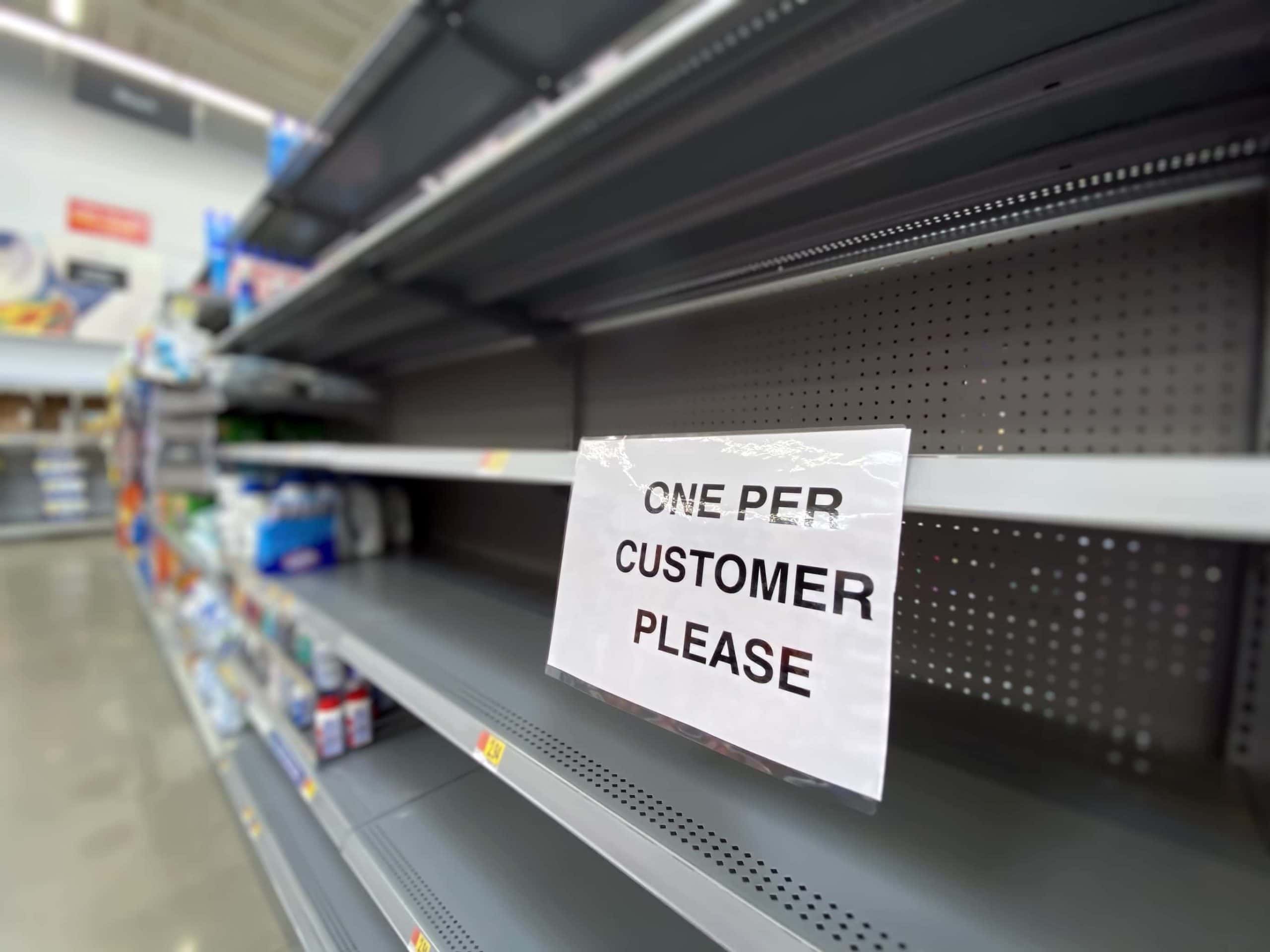 Ein Zettel mit der Aufschrift: „One per Customer“, der an einem leeren Supermarktregal hängt.