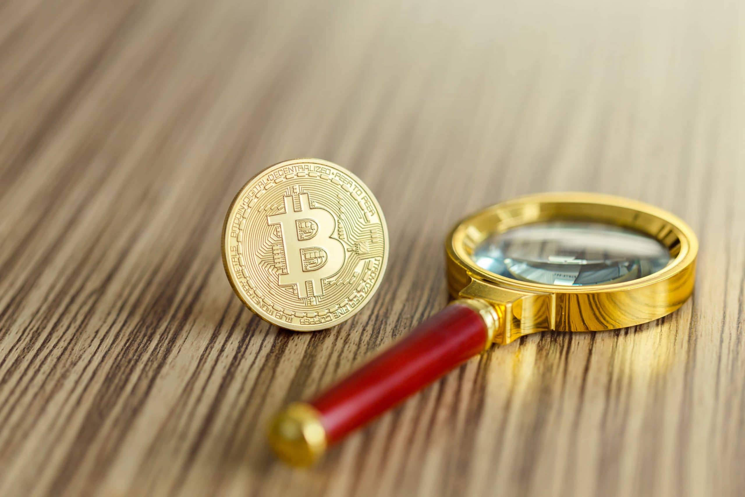 Be your own Analyst: Das On-chain-Verhalten von Bitcoin Eine Bitcoin-Münze steht neben einer Lupe auf einem Holztisch