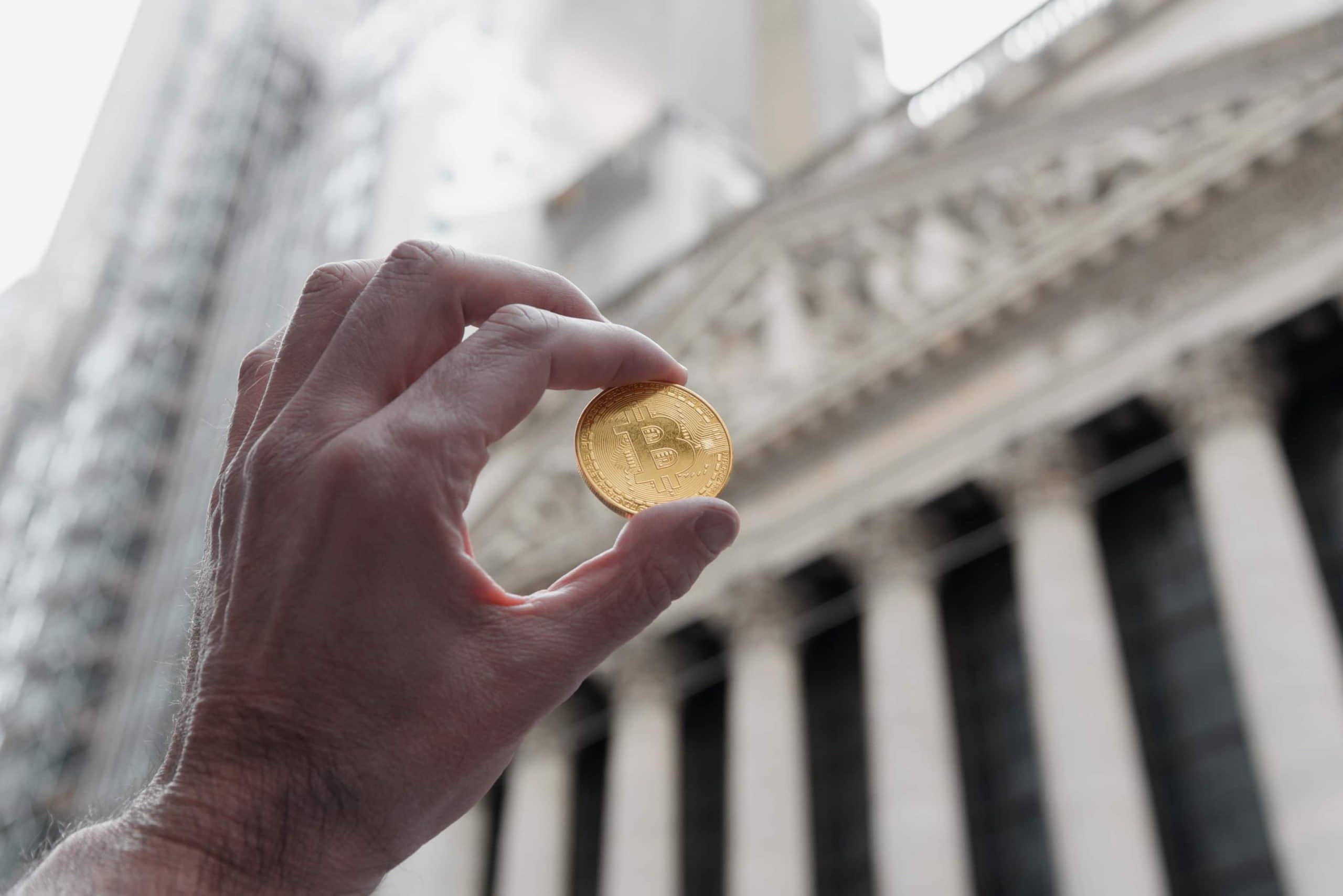 Ein Mann hält eine Bitcoin-Münze, im Hintergrund der New Yorker Börse in der Wall Street