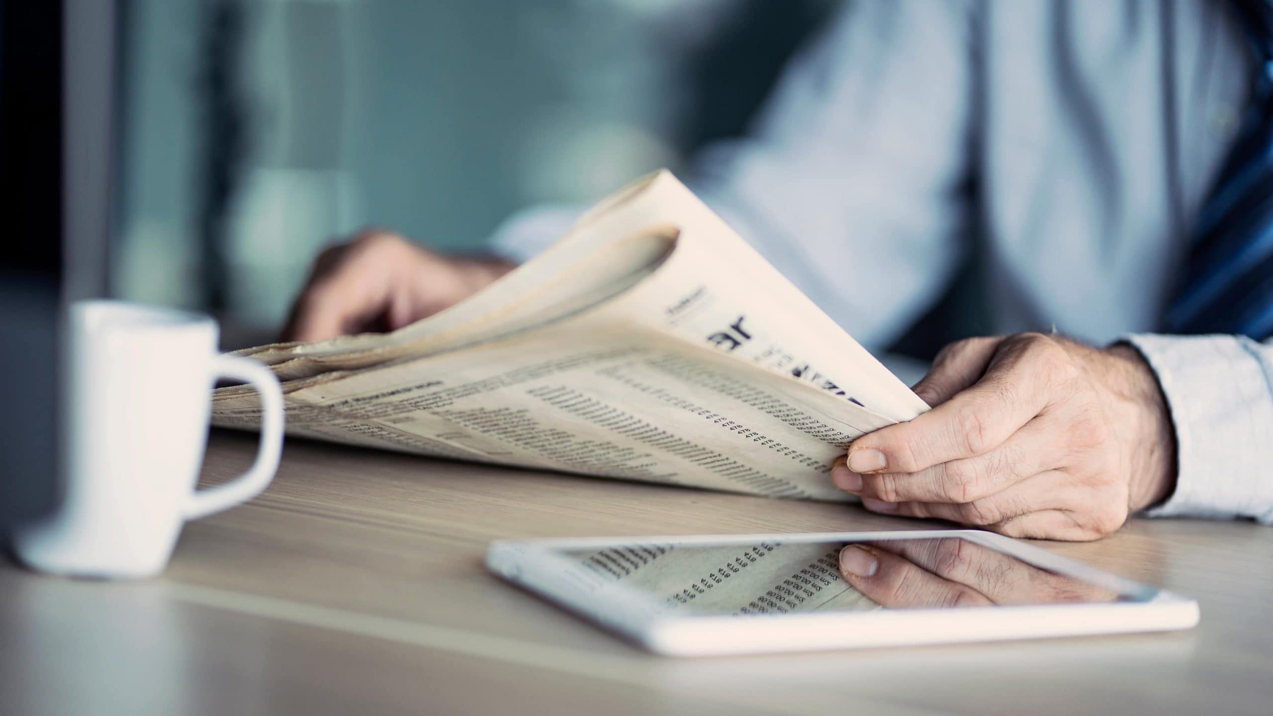 Geschägtsmann sitzt vor einem hölzernen Tisch, auf dem eine weiße Tasse steht und ein Tablet liegt und ließt Zeitung