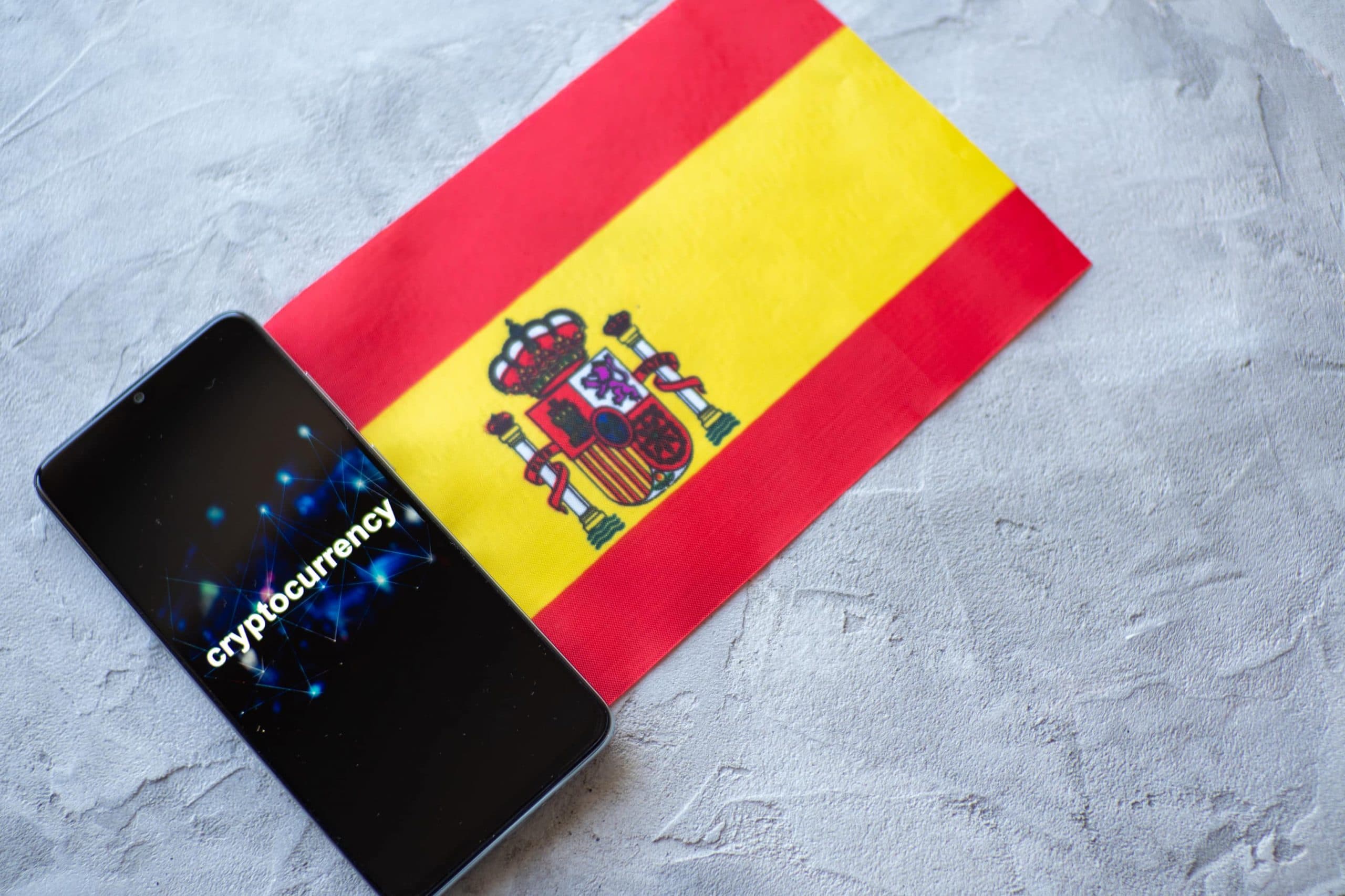 Ein Smartphone auf dem Kryptowährung auf Englisch steht, liegt neben einer ausgebreiteten spanischen Flagge, die auf einem steinernen Untergrund liegt.