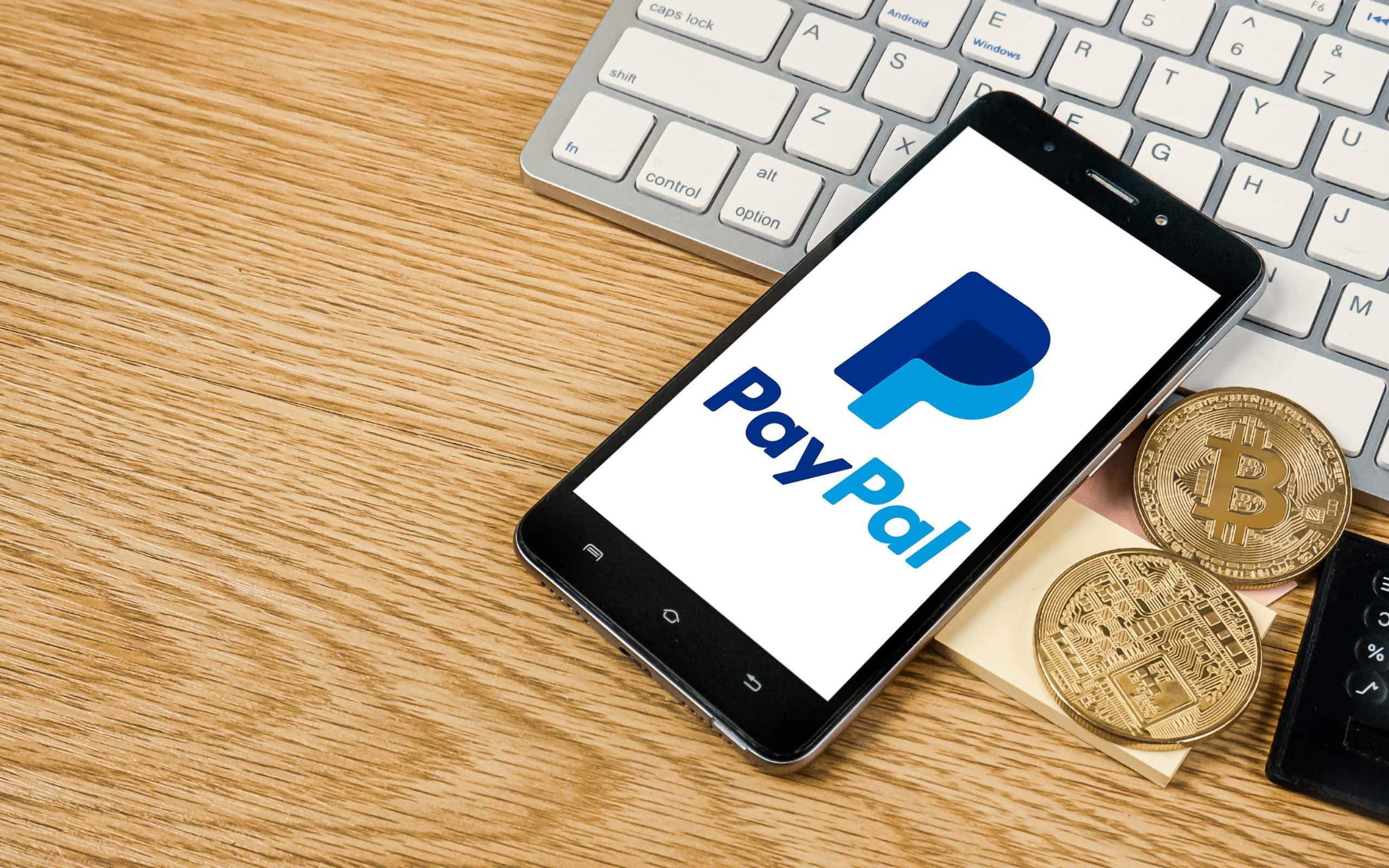 Ein Smartphone, auf dem das PayPal-Logo zu sehen ist, liegt angelehnt an eine Computertastatur auf einem Holztisch. Daneben liegen zwei Bitcoin-Münzen