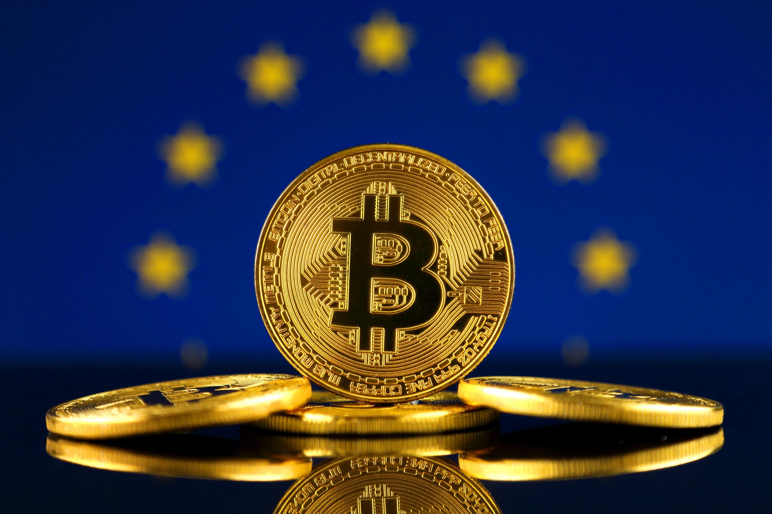 Im Vordergrund sind drei goldene Bitcoin-Münzen zu sehen. Im Hintergrund ist die Flagge der europäischen Union.