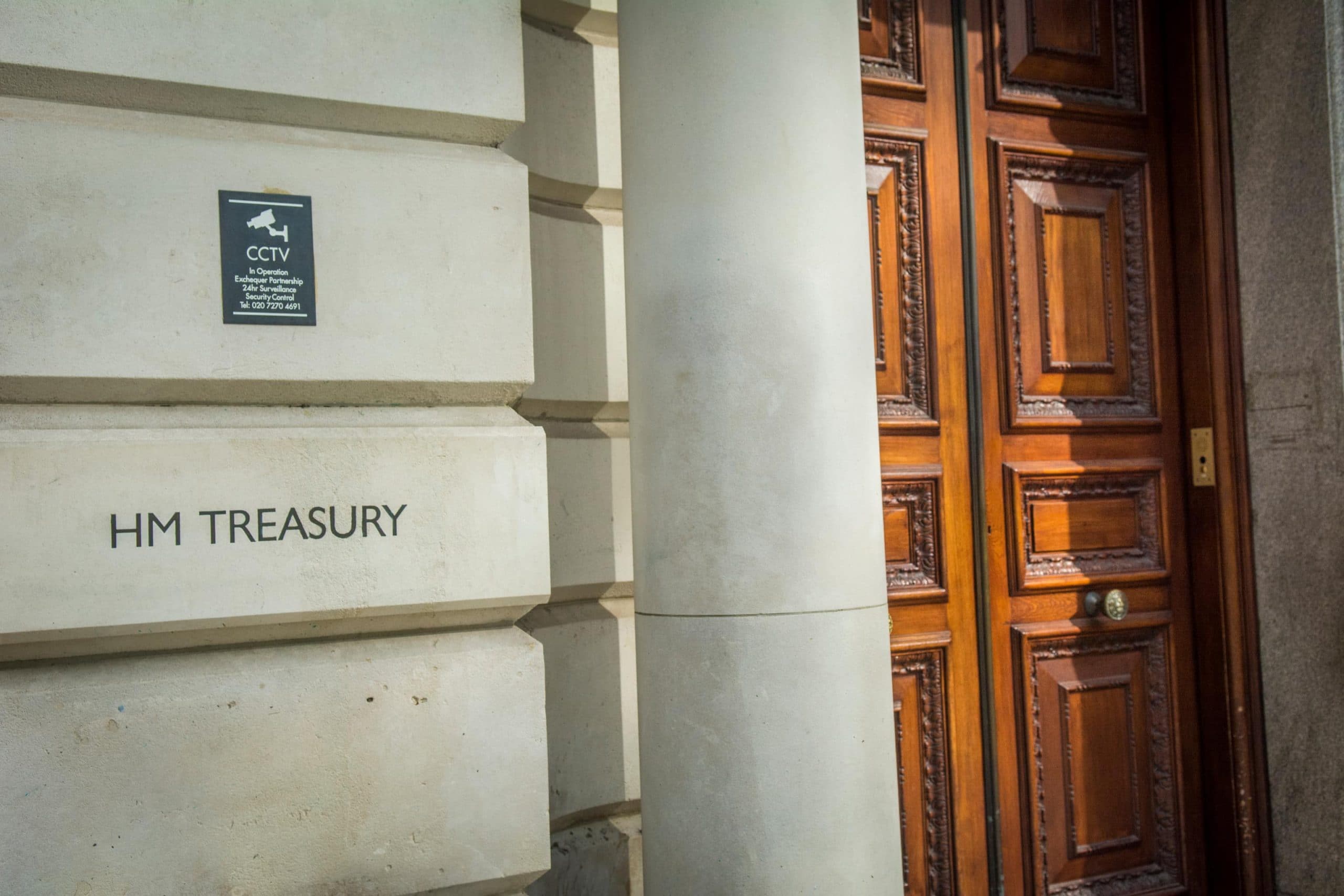 Das Logo der HM Treasury ist auf einer Steinwand zu sehen. Daneben ist eine Steinsäule und eine große, massive Holztür zu sehen.