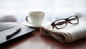 Ein Laptop, auf dem ein Kugelschreiber liegt, eine Kaffeetasse und eine Zeitung, auf der eine Brille liegt, liegen auf einem Holztisch.