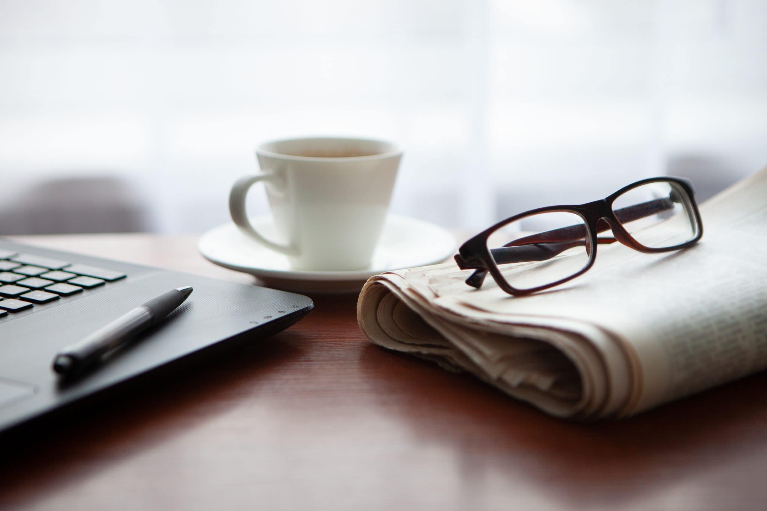 Ein Laptop, auf dem ein Kugelschreiber liegt, eine Kaffeetasse und eine Zeitung, auf der eine Brille liegt, liegen auf einem Holztisch.