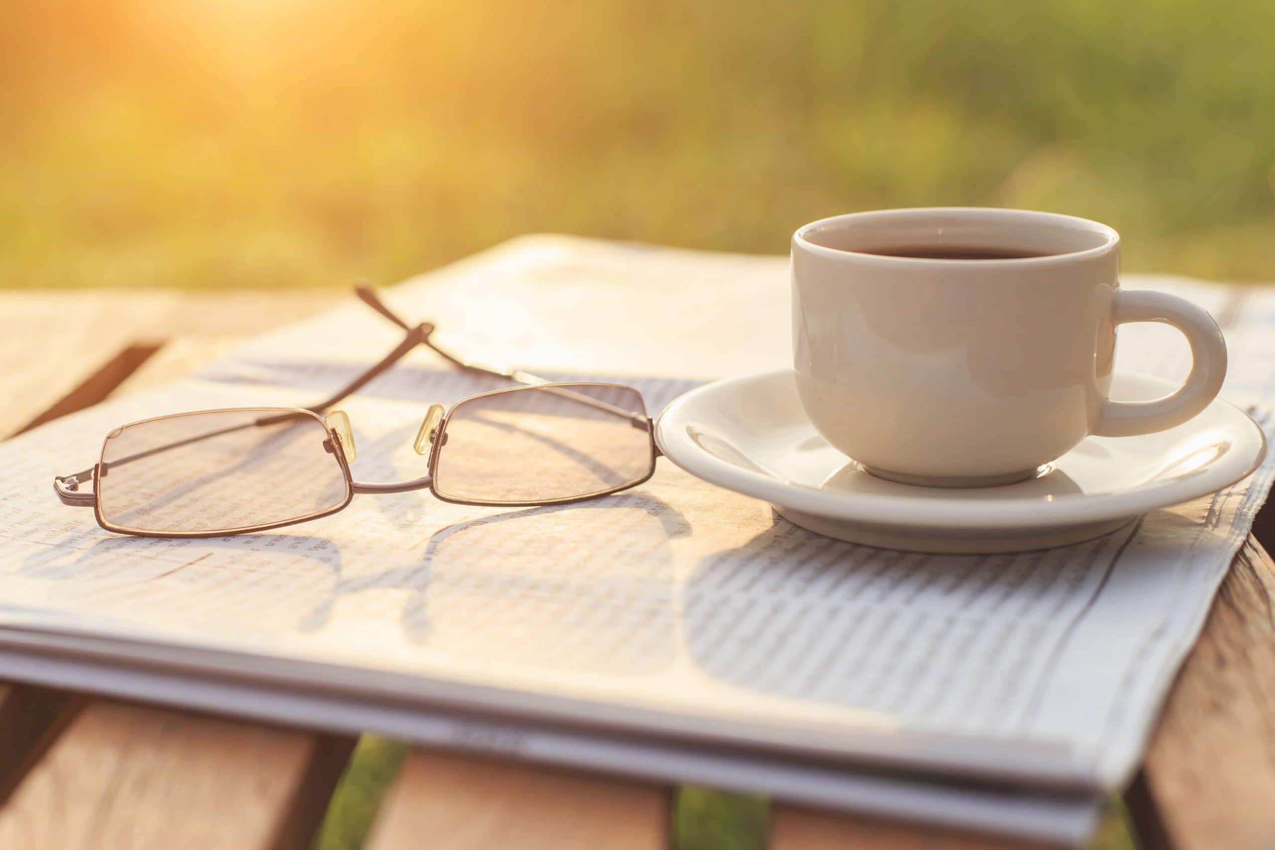 Brille und Kaffeetasse stehen auf einer Zeitung, die auf einem Holztisch liegt.