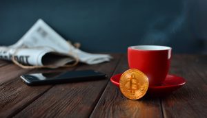 Eine Zeitung, ein schwarzes Smartphone, eine rote Kaffeetasse mit Untersetzer und eine Bitcoin-Münze stehen auf einem Holztisch.