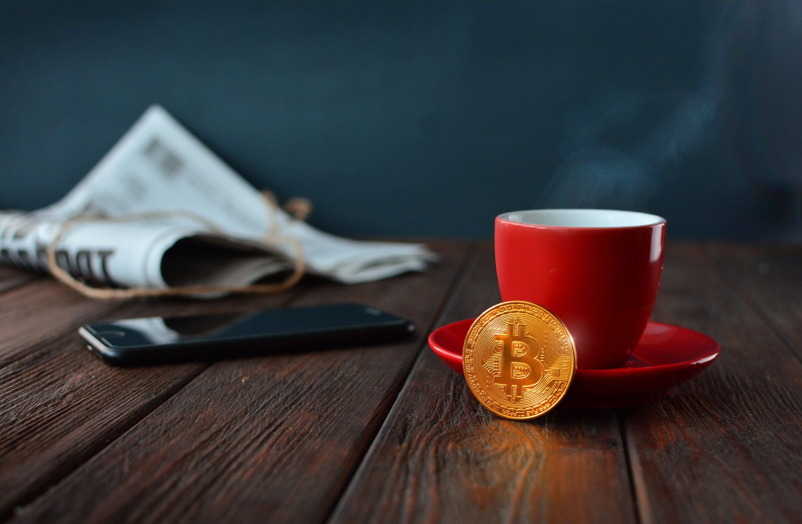 Eine Zeitung, ein schwarzes Smartphone, eine rote Kaffeetasse mit Untersetzer und eine Bitcoin-Münze stehen auf einem Holztisch.