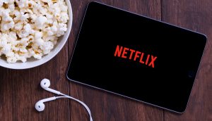 Das Logo von Netflix ist auch einem Smartphone zu sehen. Daneben steht Popcorn und darunter sind weiße Kopfhörer zu sehen.