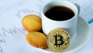 Kaffe-Tasse mit Gebäck und einem Bitcoin auf der Untertasse steht auf einer Unterlage, die aus Kurs-Diagrammen besteht.