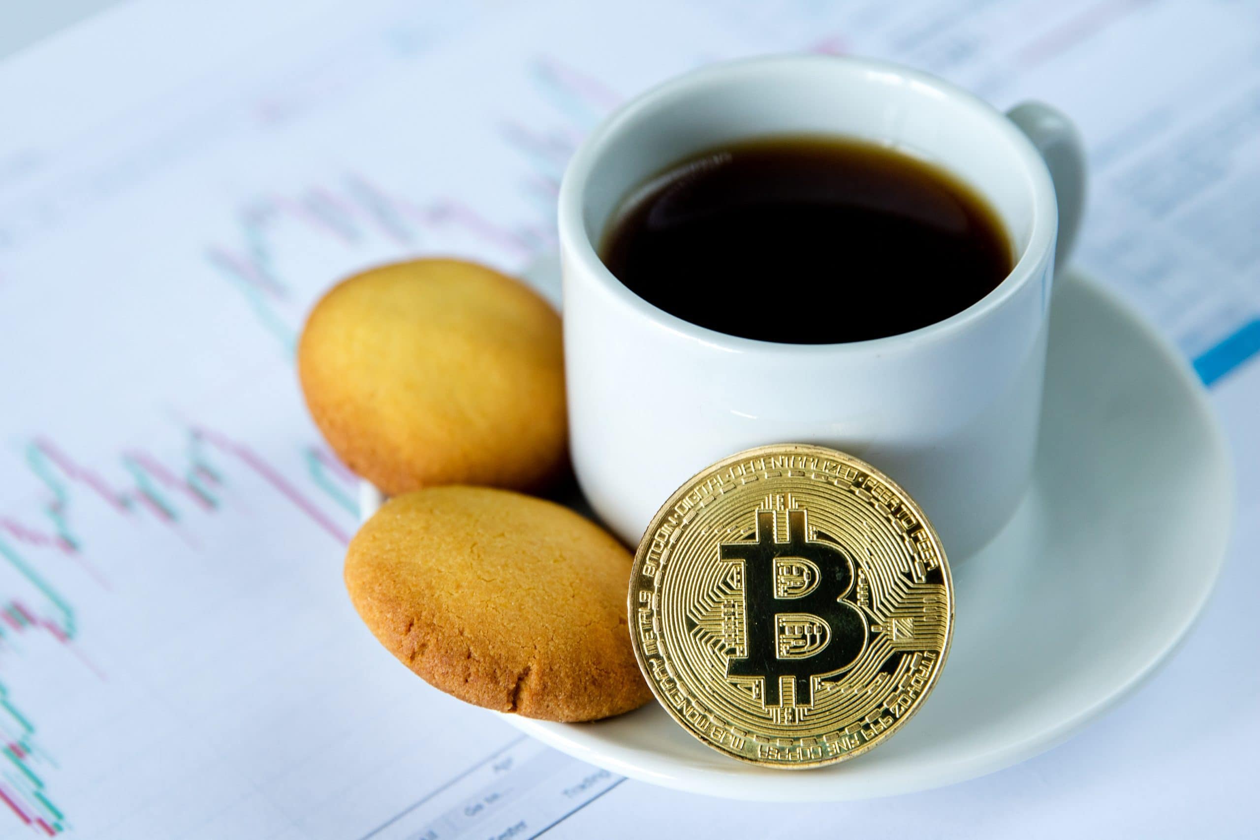 Kaffe-Tasse mit Gebäck und einem Bitcoin auf der Untertasse steht auf einer Unterlage, die aus Kurs-Diagrammen besteht.