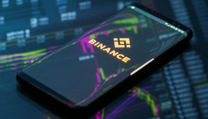 Ein Smartphone liegt auf einem Computerbildschirm, der Aktienkurse anzeigt. Auf dem Bildschirm des Smartphones ist das Logo von Binance zu sehen.