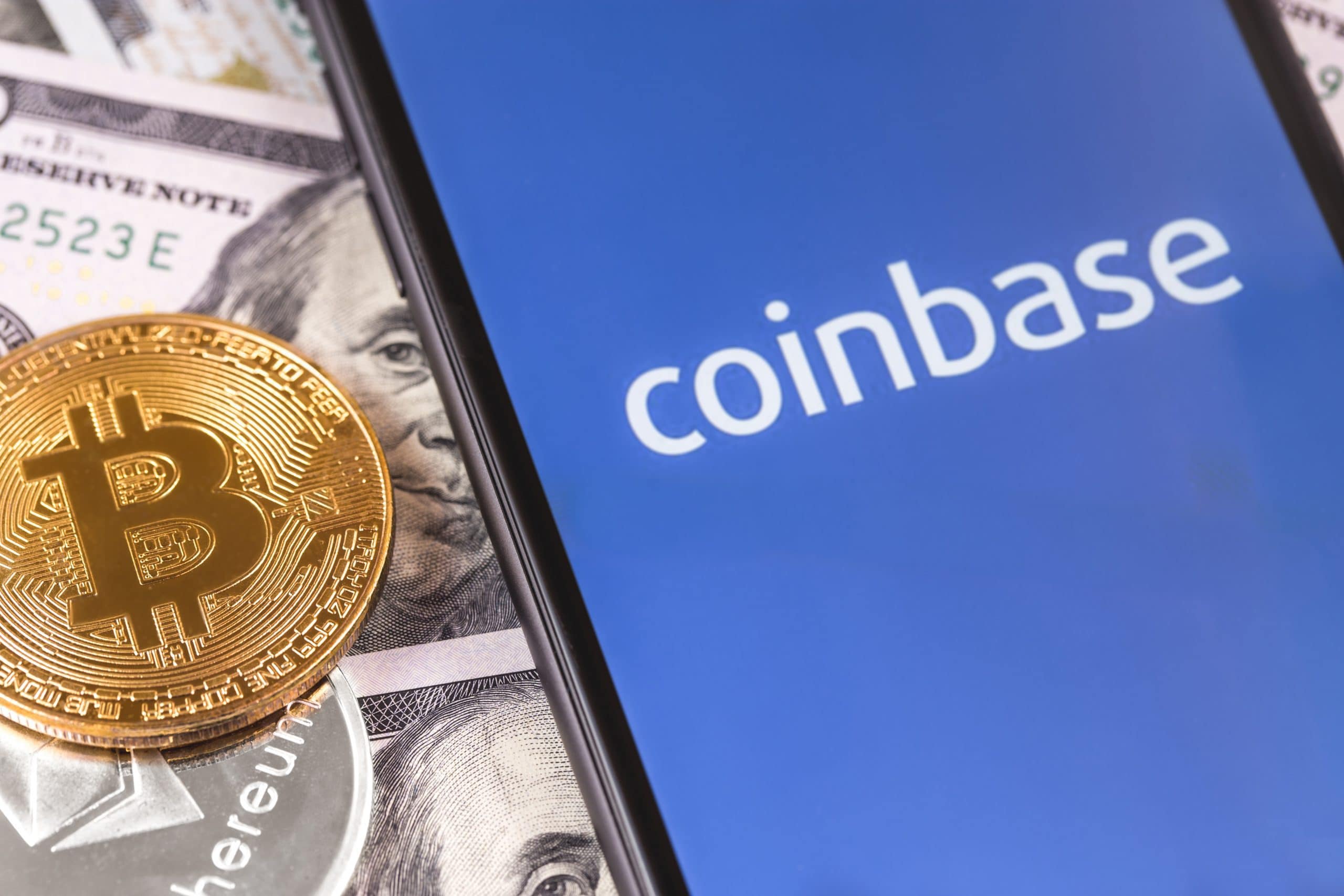 Ein Smartphone-Bildschirm zeigt das Coinbase-Logo. Daneben liegen US-Dollar-Noten und eine Bitcoin-Münze.