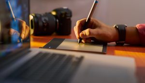 Eine Hand zeichnet etwas auf ein Tablet. daneben steht ein MacBook.