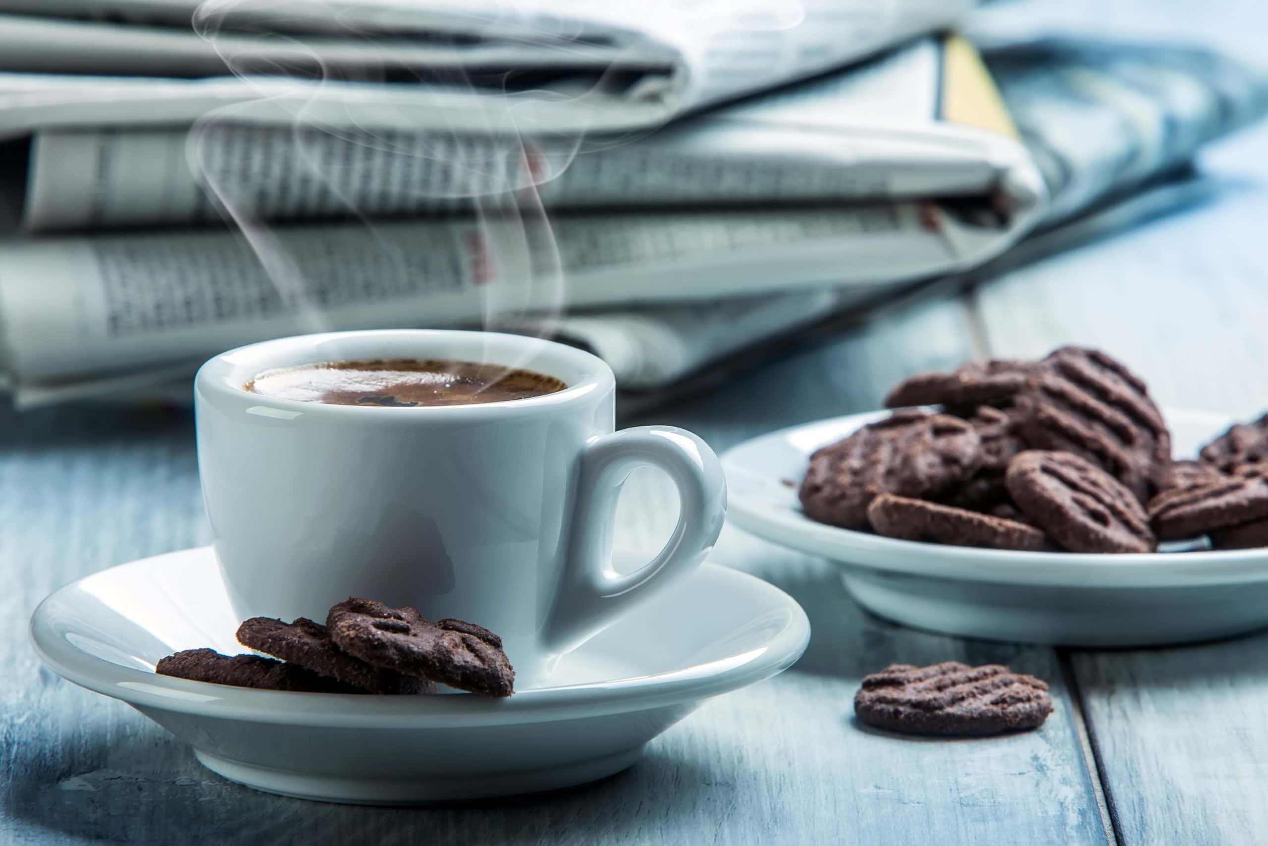 Eine weiße Kaffeetasse samt Untersetzer steht auf einem Tisch. Daneben steht ein Teller mit Gebäck. Dahinter liegt ein Stapel Zeitungen.